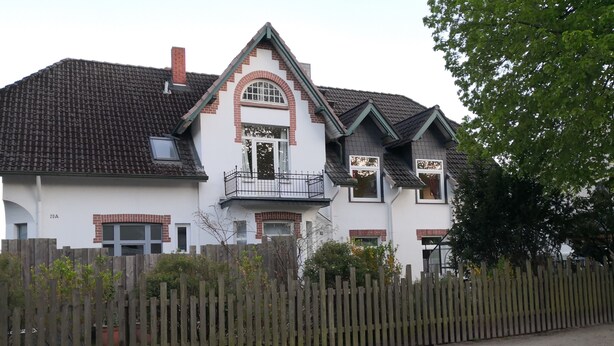 A two-story house with a balcony, a brick chimney, and a wooden fence. - Vacation apartment in a beautiful location between 2 lakes for 2-6 people (Malente)