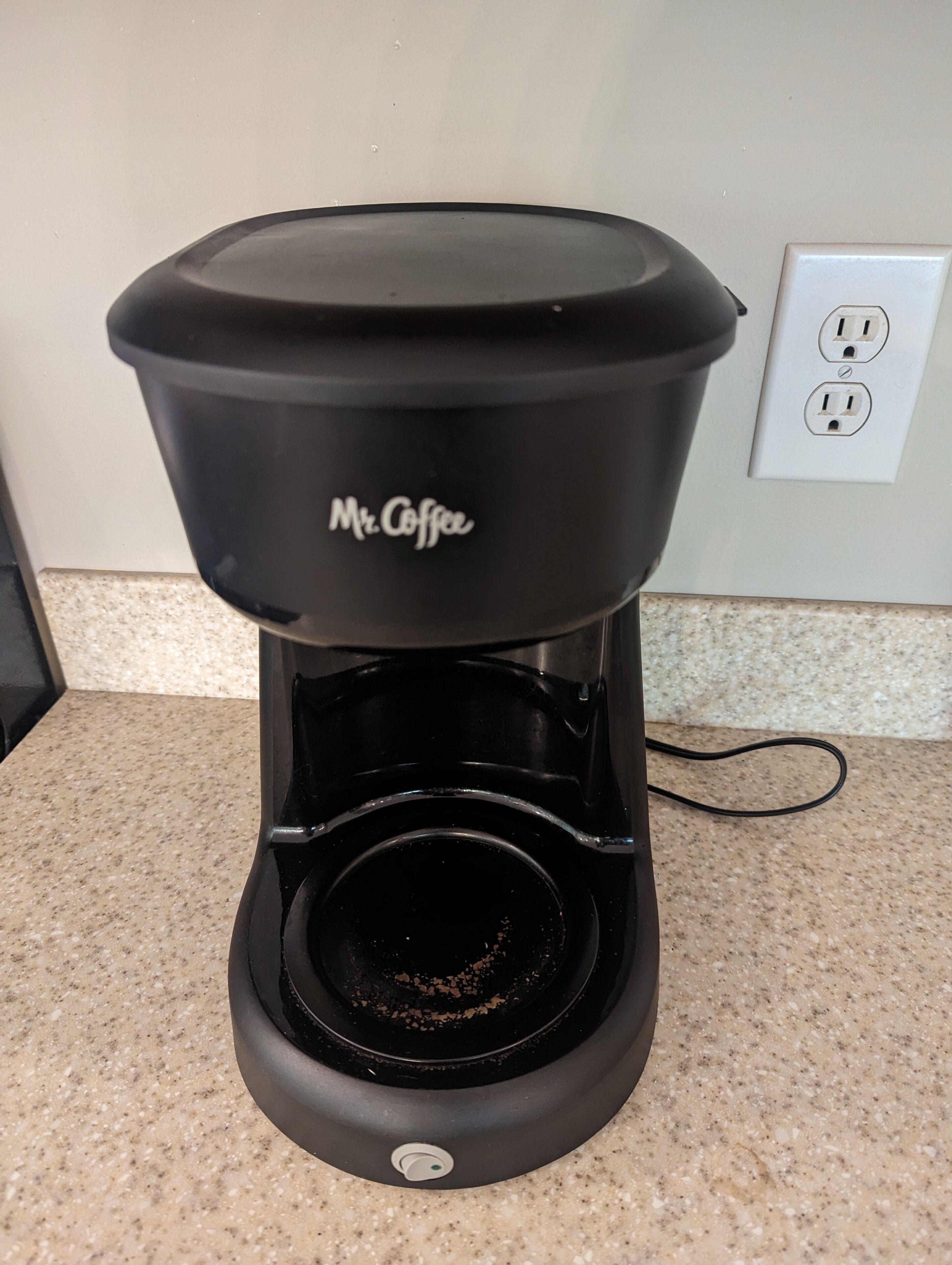 A black Mr. Coffee coffee maker on a kitchen counter with a granite backsplash and a power outlet in the background.