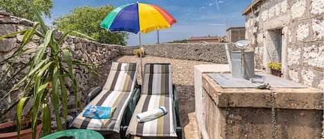 A stone wall, two striped lounge chairs, a colorful umbrella, a metal bucket, and a green table.