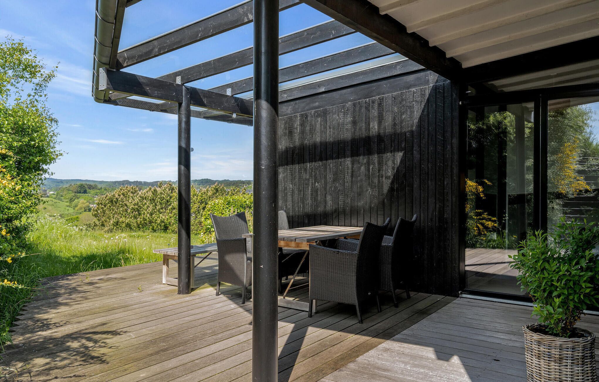 Une terrasse extérieure avec une table et des chaises, entourée d’une pergola en bois et de murs en verre offrant une vue sur la campagne.
