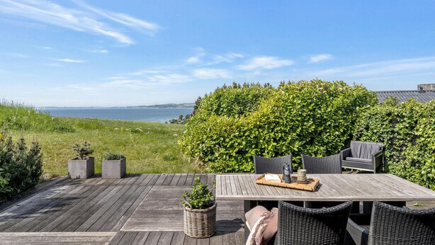 Een houten terras met een tafel en stoelen, uitkijkend op een waterpartij en een groene haag.