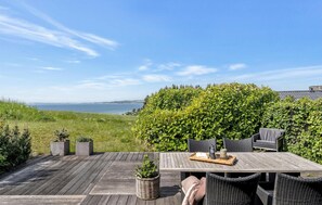 Una terraza de madera con una mesa y sillas, con vista a un cuerpo de agua y un seto verde.