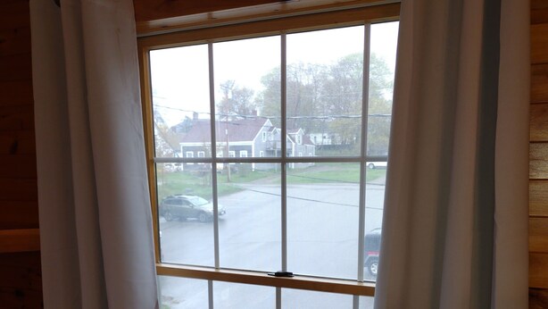 A window with white curtains, a view of a parking lot, and a building in the background. - Eastern Beacon Inn Room 3 (Lubec)