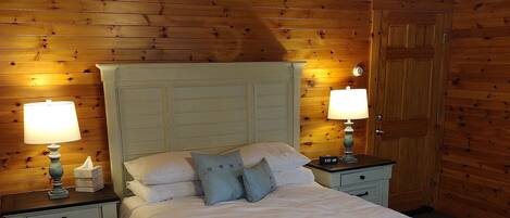 A bedroom with a wooden ceiling, a bed with white bedding, two bedside tables with lamps, and a wooden door.