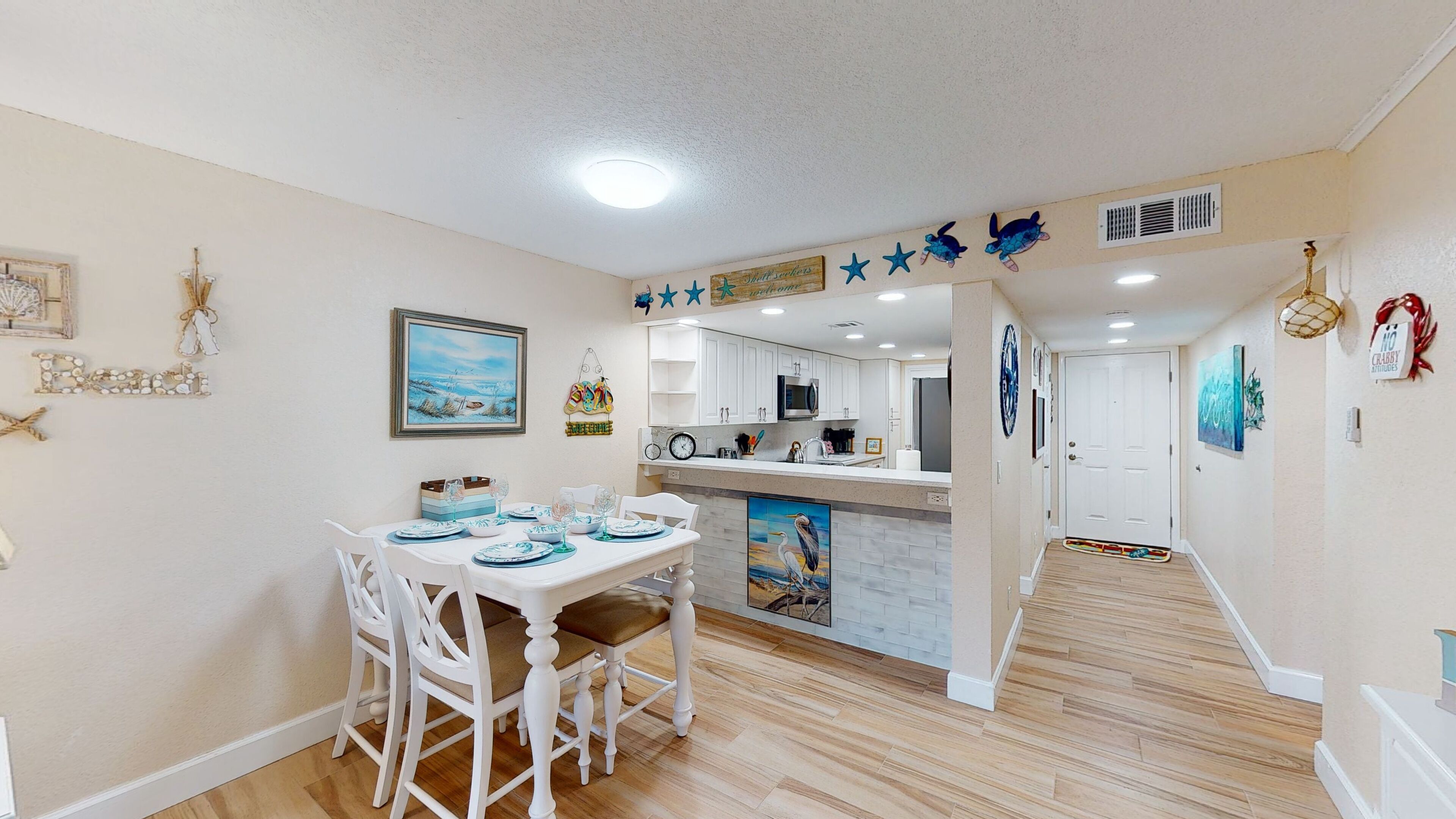 A dining area with a table set for four, white chairs, a kitchen in the background, and a hallway leading to a door.