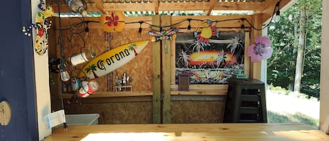 Un bar pequeño con una barra de madera, un mural de una puesta de sol y diversas botellas y vasos en estanterías.