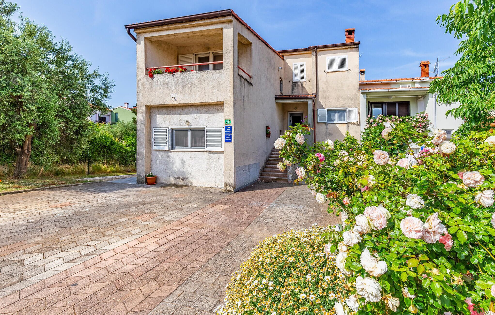 A two-story building with a balcony, a small garden with roses, and a paved driveway.