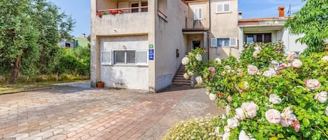 Ein zweistöckiges Gebäude mit Balkon, einem kleinen Rosengarten und einem gepflasterten Auffahrt.