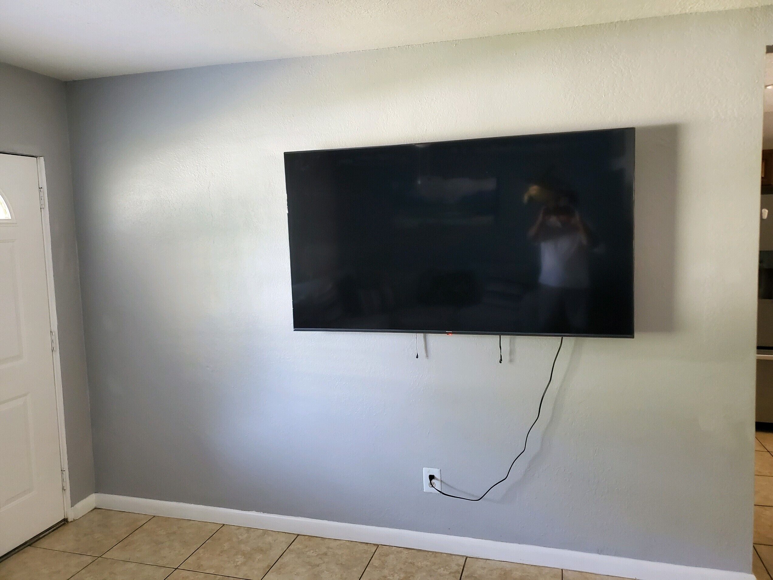 A room with a flat-screen TV mounted on a light grey wall, a white door, and a tiled floor.