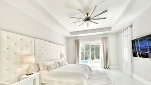Un dormitorio moderno con cabecera capitonada, cama grande, ventilador de techo y vista a una piscina a través de puertas de cristal.