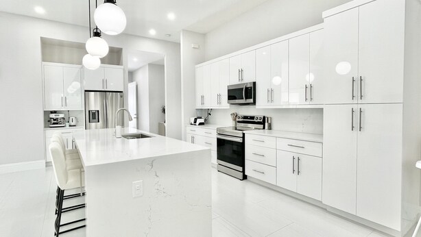 Una cocina moderna con armarios blancos, una isla central, electrodomésticos de acero inoxidable e iluminación colgante.