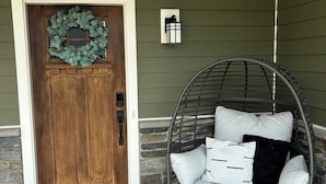 Une porte en bois ornée d’une guirlande, un mur en pierre, un fauteuil suspendu avec des coussins et un luminaire fixé au mur.