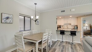 A dining area with a wooden table and white chairs, a kitchen with white cabinets, and a gray sofa in the living area. - Newly Renovated 2BR/2BA Flat/3 Smart TV’s/WIFI/Pool & Tennis/Pickleball Views (Hilton Head Island)