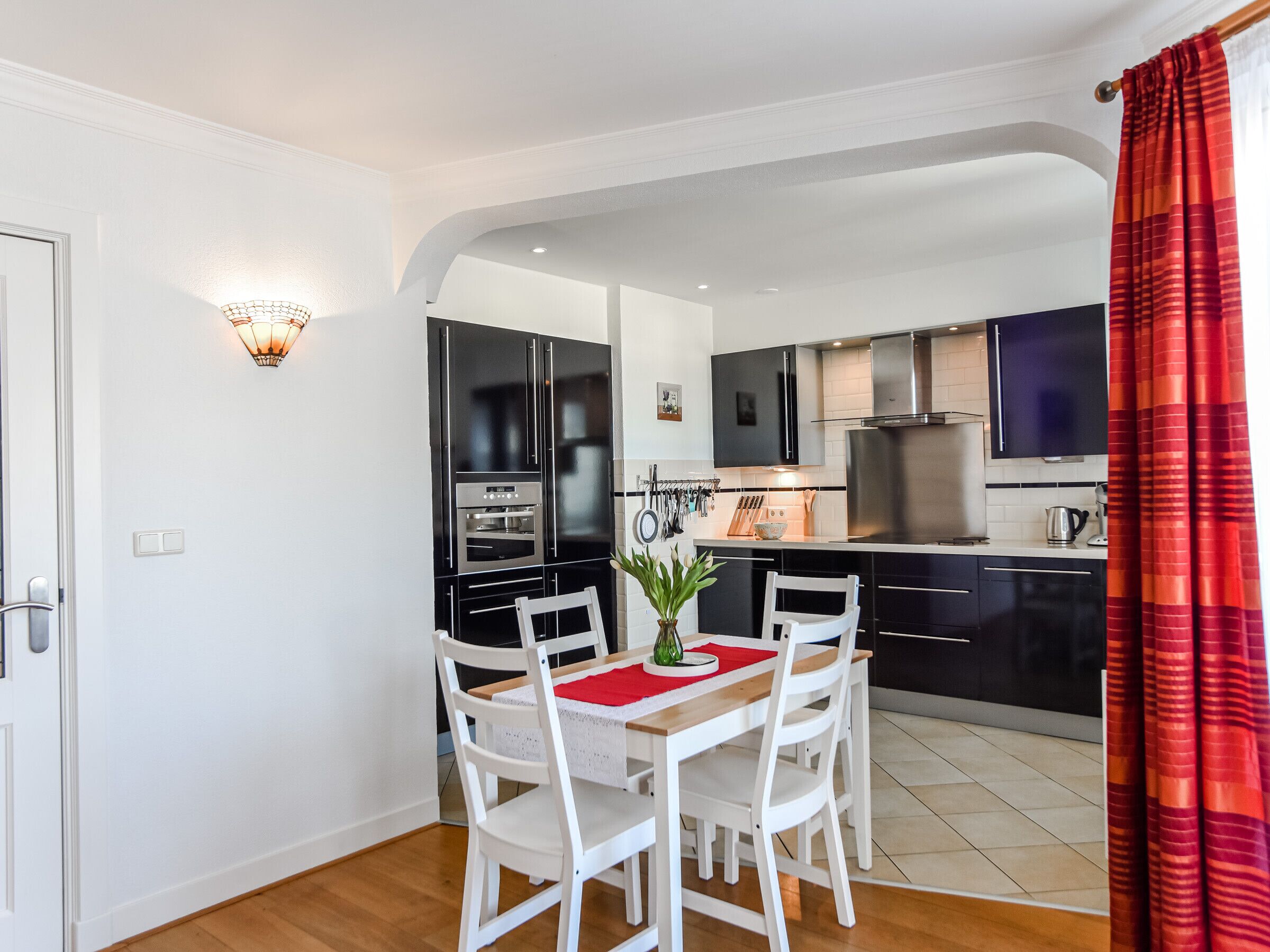 Une cuisine compacte dotée d’appareils électroménagers modernes, un coin repas avec une table et des chaises blanches, et un rideau rouge.