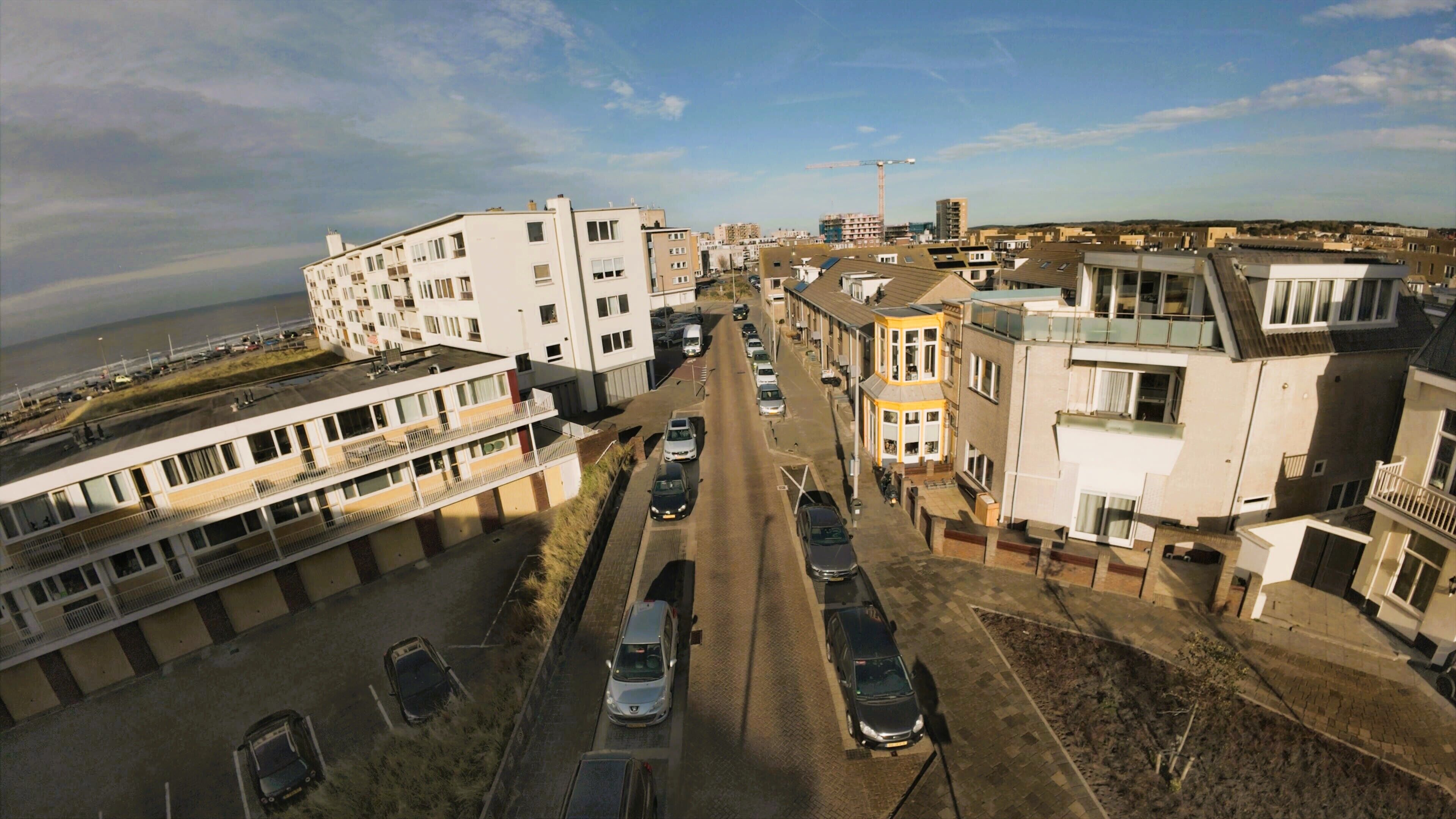 Vue aérienne d’une rue résidentielle bordée d’immeubles à plusieurs étages, avec des voitures garées et une grue de construction visible au loin.