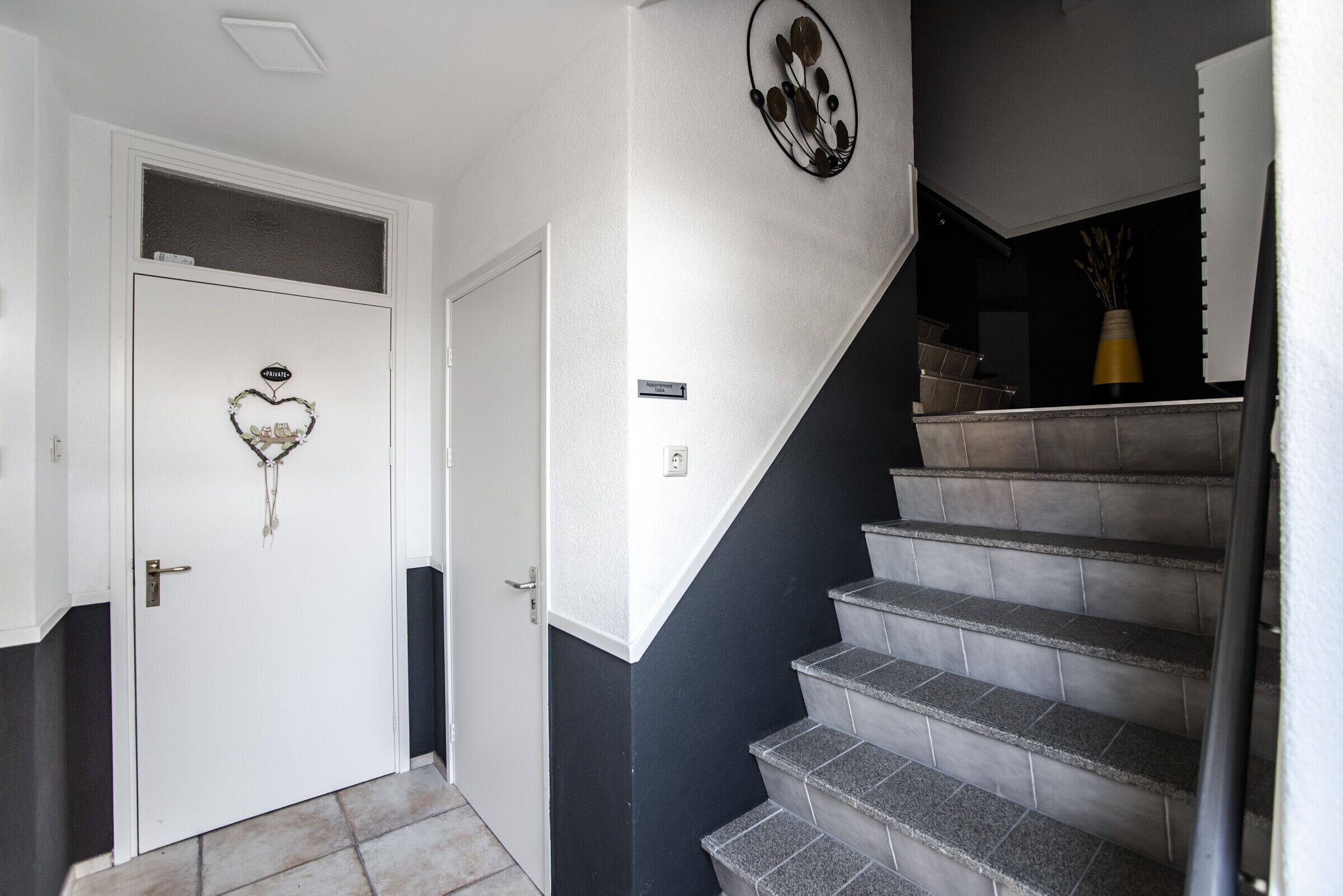 Una escalera con pared negra y peldaños grises, una puerta blanca con adorno de corazón y un cartel en la pared.