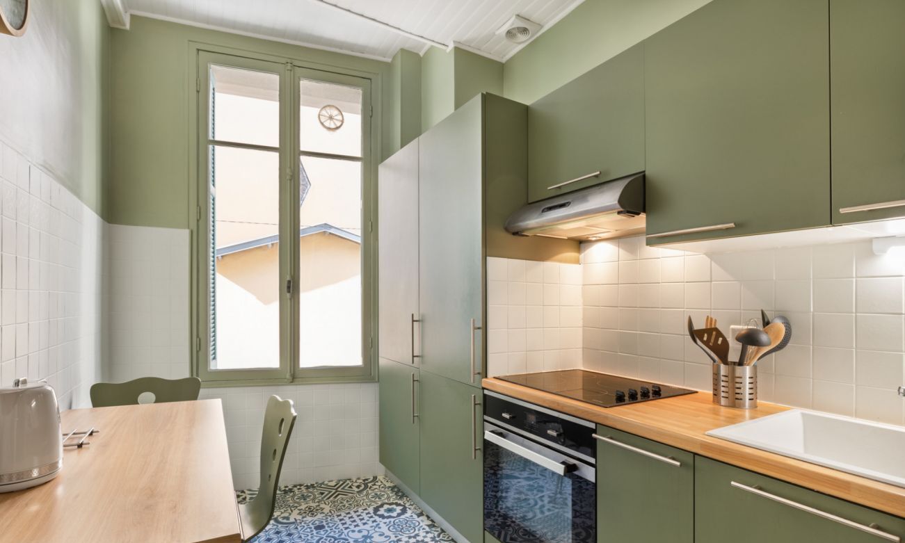 Een compacte keuken met groene kastjes, een houten aanrechtblad en een raam met uitzicht op een huis.