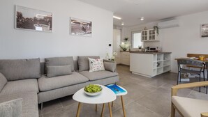 A modern living room with a grey sofa, a small white table with a bowl of fruit, and a dining area with a wooden table and chairs. - Beach front apartment in Mali Losinj with WiFi (Mali Losinj)