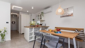 A modern kitchen with white cabinetry, a wooden dining table, and a bench. There is a hanging light fixture above the table and a framed picture on the wall. - Beach front apartment in Mali Losinj with WiFi (Mali Losinj)