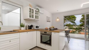 A modern kitchen with white cabinetry, a built-in oven, and a large window offering a view of a coastal town. - Beach front apartment in Mali Losinj with WiFi (Mali Losinj)
