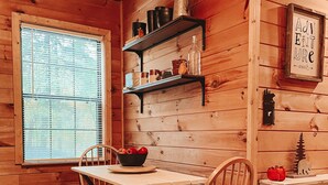 A cozy cabin interior with a wooden dining table, chairs, a bookshelf, and a window with blinds. - Mountain Cabin with creek and hiking trail! Minutes from Blue Ridge (Morganton)