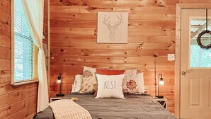 A wooden cabin bedroom with a bed, pillows, a blanket, a wall-mounted deer head artwork, and a sign indicating a hiking trail. - Mountain Cabin with creek and hiking trail! Minutes from Blue Ridge (Morganton)