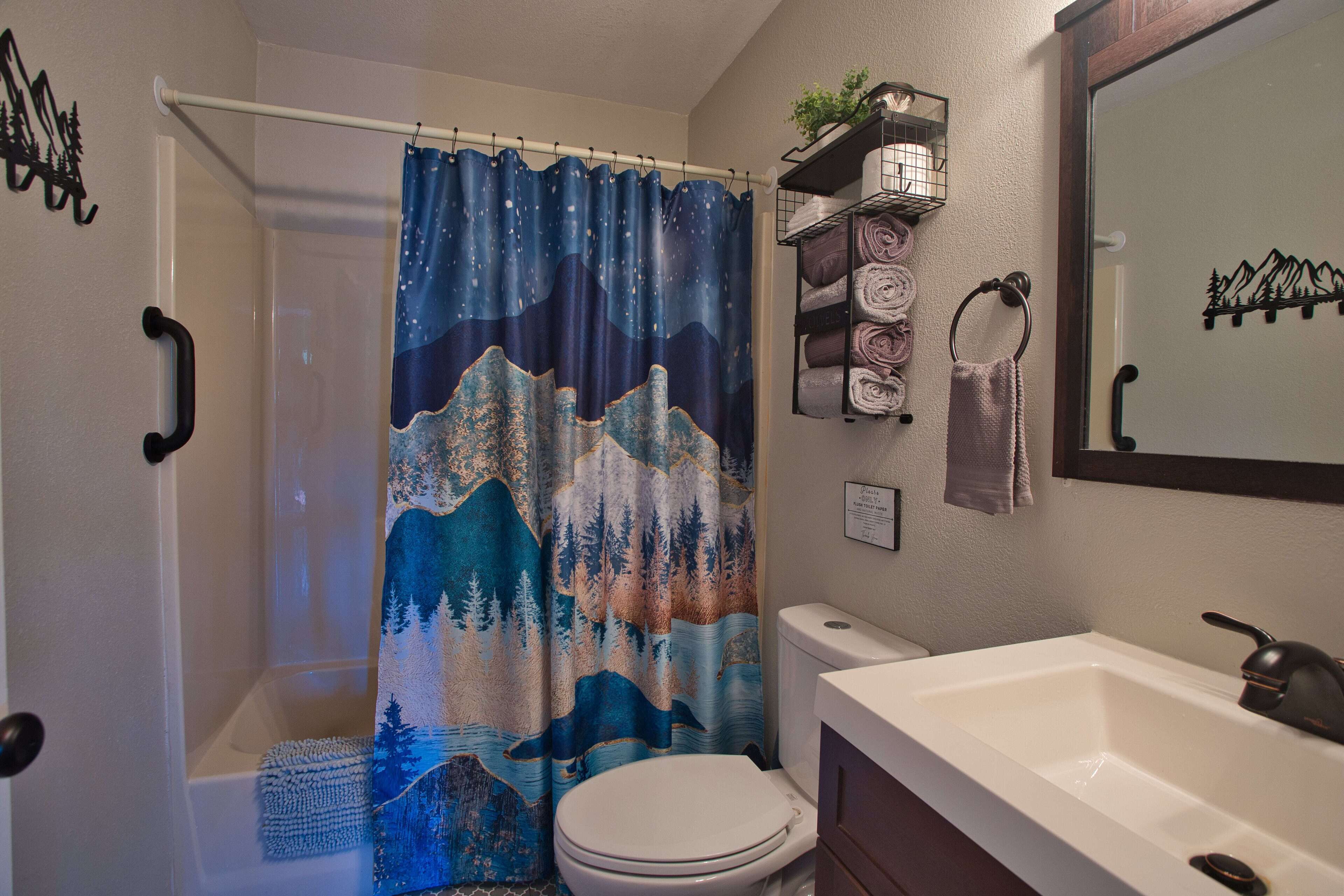 A bathroom with a shower curtain depicting mountains and trees, a toilet, a sink with a mirror, and towel racks.