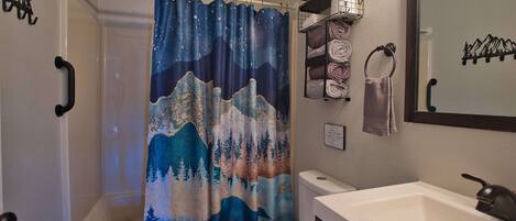 A bathroom with a shower curtain depicting mountains and trees, a toilet, a sink with a mirror, and towel racks.