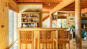 Una zona de bar con barra de madera, estante con copas y botellas, tres sillas de madera y una escalera que conduce a otra habitación.