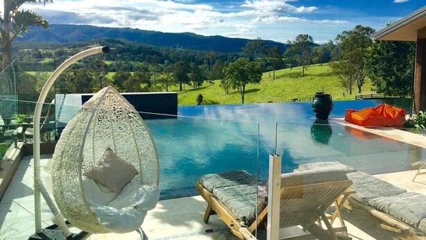 Una piscina de borde infinito con fondo de cristal, una silla colgante y sillones de descanso, rodeada de un paisaje exuberante y montañas al fondo.