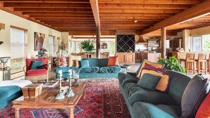 Amplia sala de estar con techo de madera alto, un sofá grande, una mesa de centro y un comedor con sillas de madera, con una cocina al fondo.