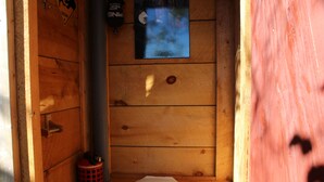 Cabina de baño de madera con inodoro blanco, un cubo de basura rojo pequeño y una ventana que da al exterior.