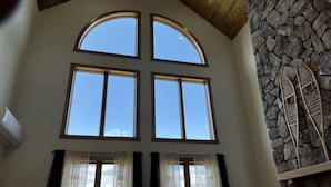 A room with a high ceiling, a stone fireplace, and four large windows with curtains. - Biwabik Bunkhouse near Giants' Ridge (Aurora)