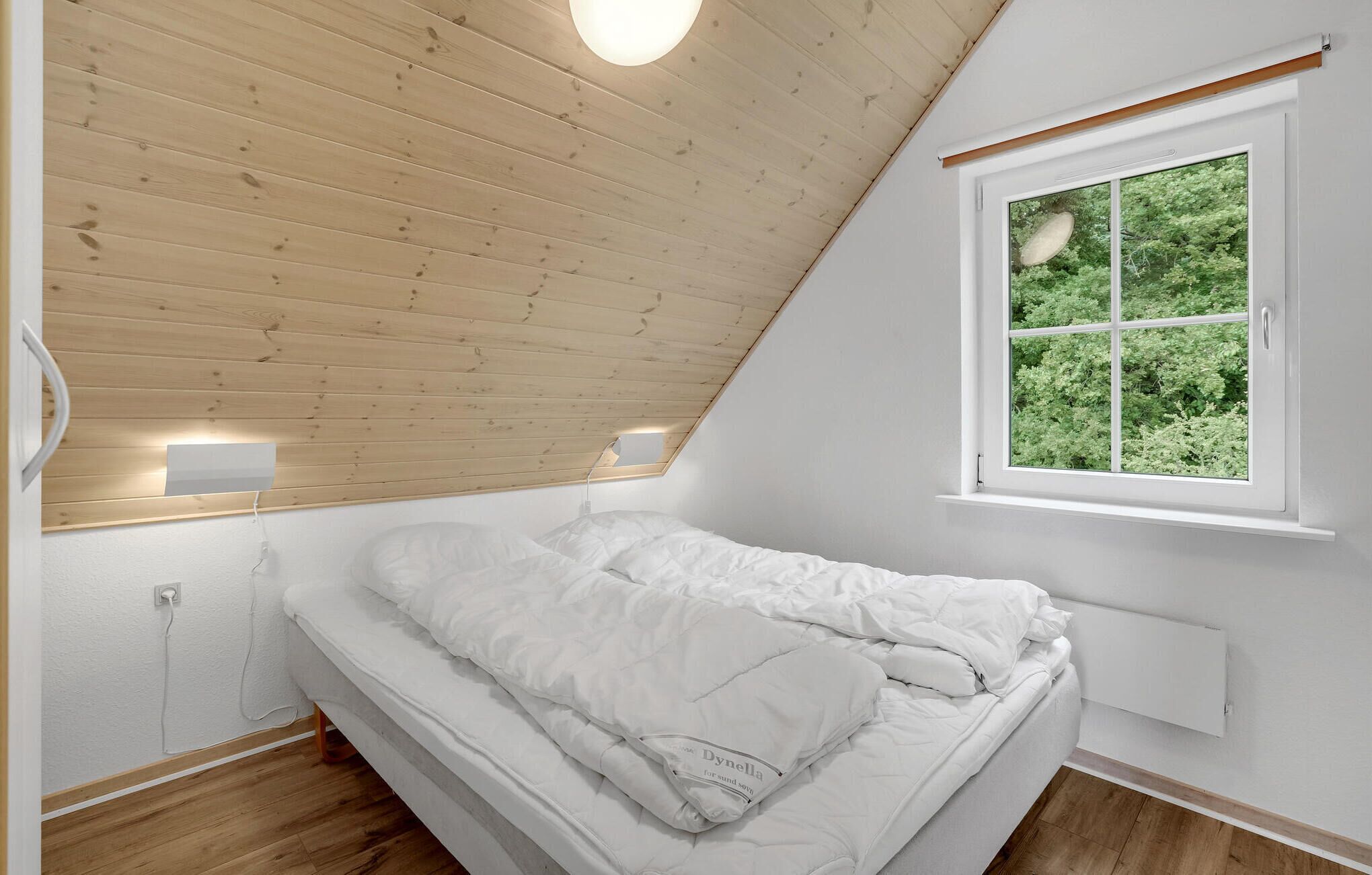 Ein Schlafzimmer mit schrägem Dach, einem Oberlicht, einem Bett mit weißer Bettwäsche und einem Fenster mit Blick auf Grünflächen.