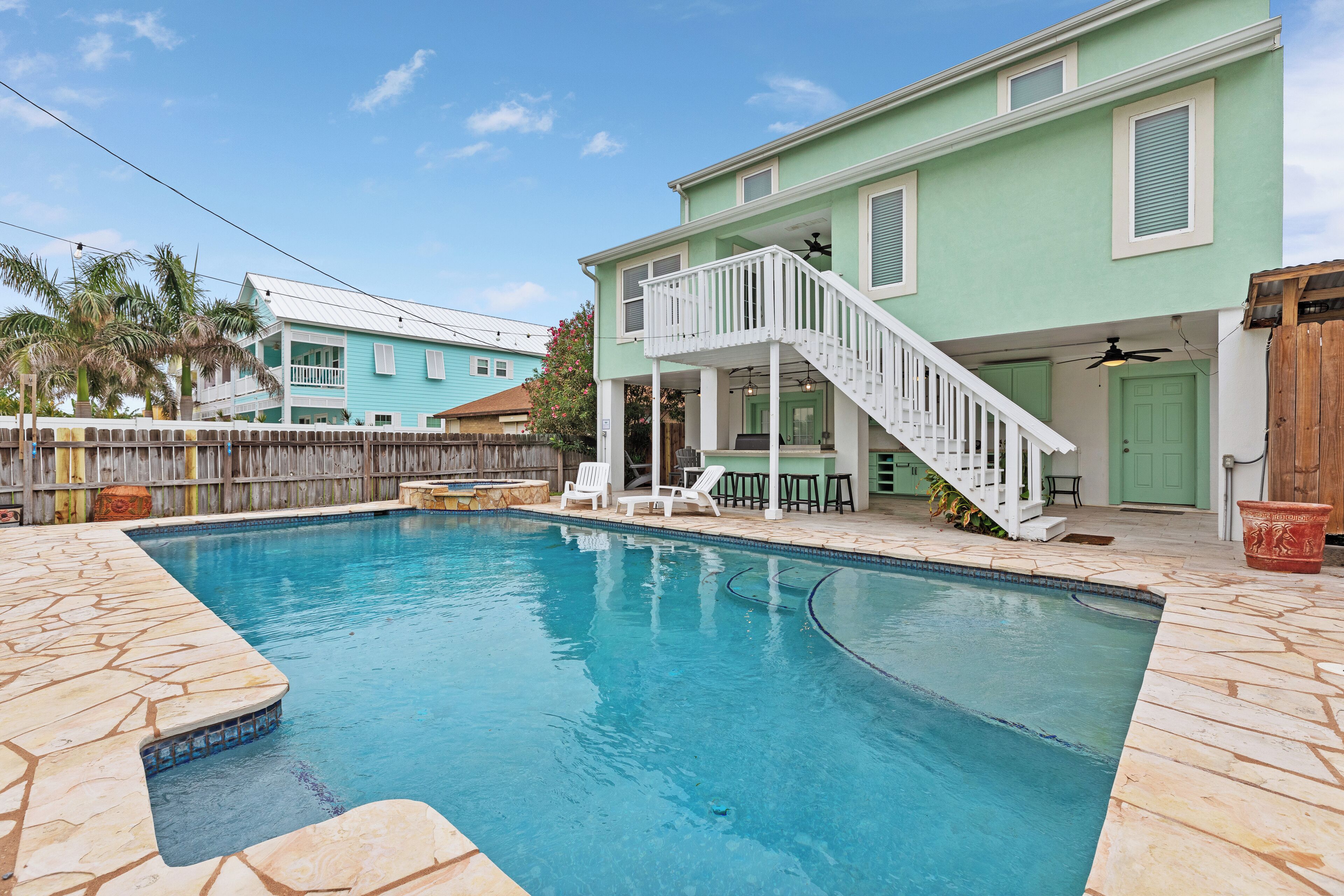 Une piscine avec une terrasse blanche, un patio couvert et une maison à deux étages dotée d’un balcon.