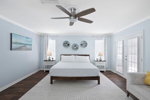 Ein Schlafzimmer mit einem Bett, Deckenventilator, einem Gemälde, Nachttischen und einem Fenster mit Jalousien.