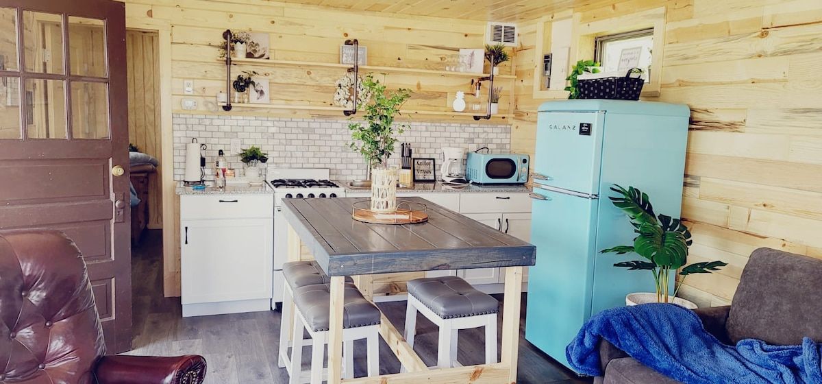 A compact kitchen with a dining table, stools, a refrigerator, and a microwave.