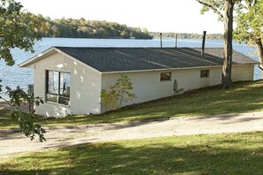 A single-story building with a sloped roof, a large window on the left, and a small door. It is situated next to a body of water with trees in the background. - Charming 2-Bedroom Lakeside Cabin with Dock Access (Alexandria)