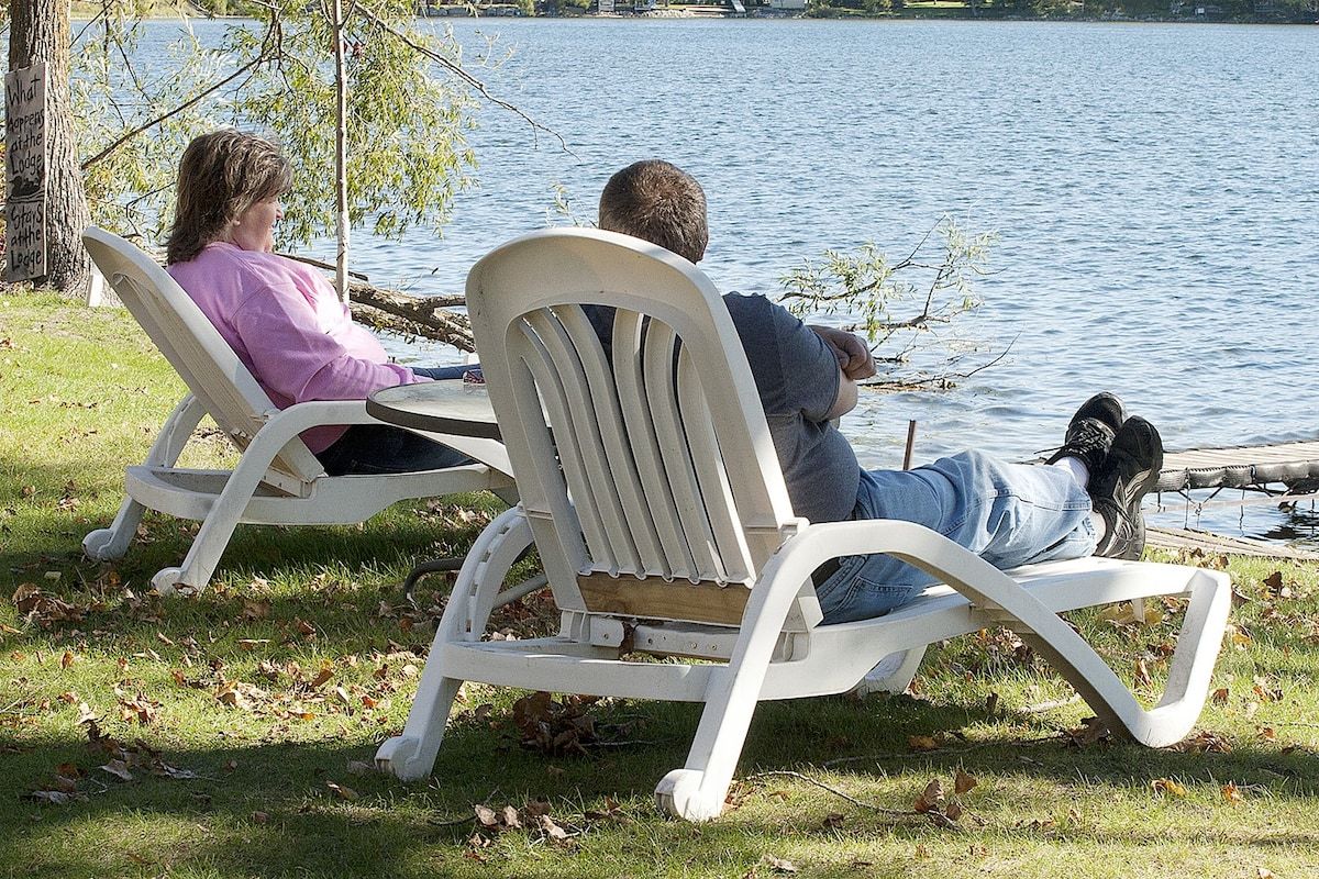 Deux personnes sont assises sur des chaises blanches, face à une étendue d’eau, et un chien est allongé sur le sol à proximité.