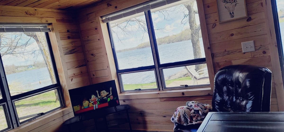 A cabin interior with wooden walls, a black leather chair, a small table, and a window overlooking a lake.