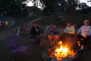 A group of people seated around a campfire in an outdoor setting with chairs and a small building in the background. - Charming 2-Bedroom Lakeside Cabin with Dock Access (Alexandria)
