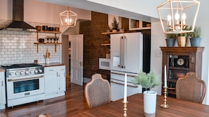 Eine moderne Küche mit weißen Schränken, einem eingebauten Herd und einer Wand mit Backsteinverkleidung. Es gibt einen Essbereich mit einem Holztisch und Stühlen sowie eine darüber hängende Kristallleuchter.