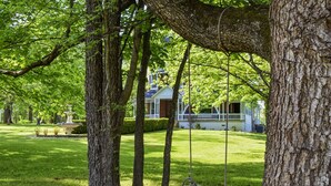 Eine Holzhängematte hängt an einem großen Baum in einem gepflegten Garten, im Hintergrund ist ein Haus zu sehen.