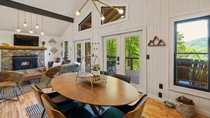 A spacious living room with a fireplace, a round wooden table, and comfortable seating. There are multiple windows offering views of the outdoors.