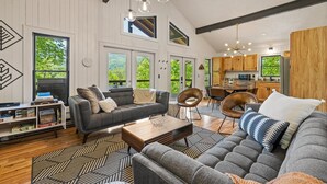 A spacious living room with a gray sectional sofa, a wooden coffee table, and a black and white patterned rug. There are multiple windows providing natural light and views of the outdoors.