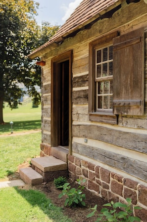 A rustic log cabin with a wooden door and window, stone foundation, and steps leading to the entrance.
