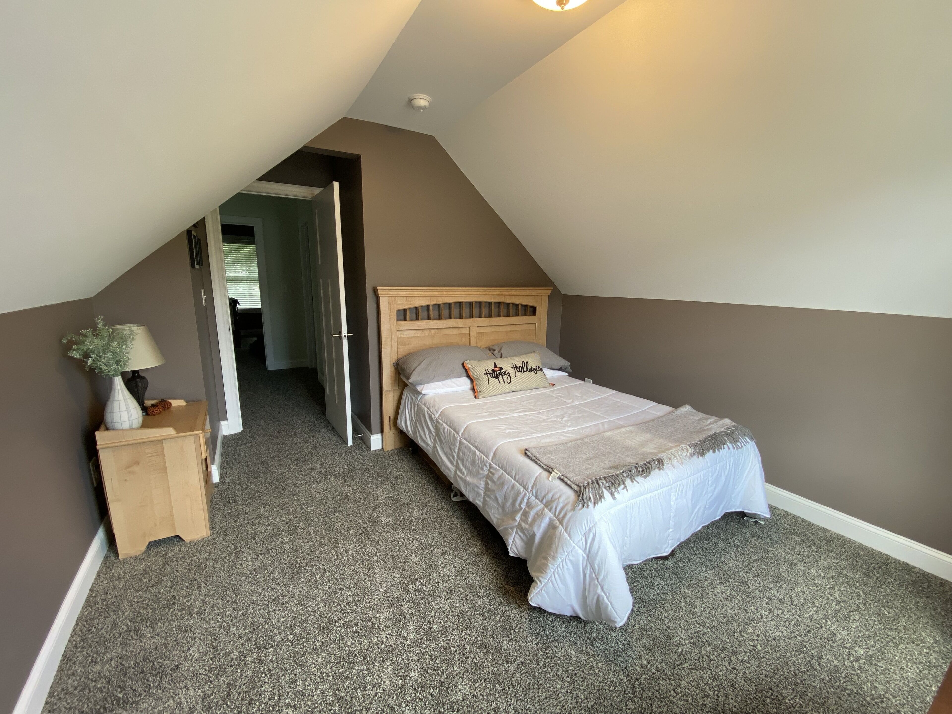 A bedroom with a sloped ceiling, a bed with a headboard, a nightstand with a lamp, and a door leading to another room.