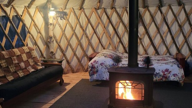 A cozy interior of a yurt with a central wood-burning stove, a sofa, and a bed with floral bedding.