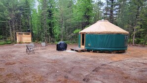 A yurt with a green exterior and a beige roof, surrounded by a forest. There is a small wooden structure and a black object, possibly a storage container, near the yurt.
