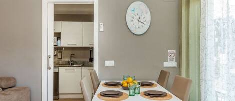 Un comedor con una mesa blanca dispuesta para cuatro personas, un reloj en la pared y una cocina al fondo.
